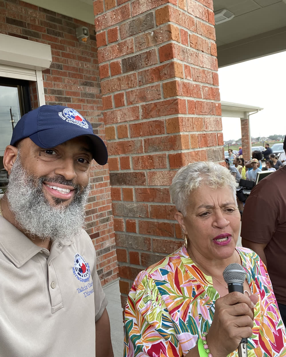 Tahir Charles in a Harris County WCID 89 cap and polo beside a speaker at the microphone during an outdoor district event.