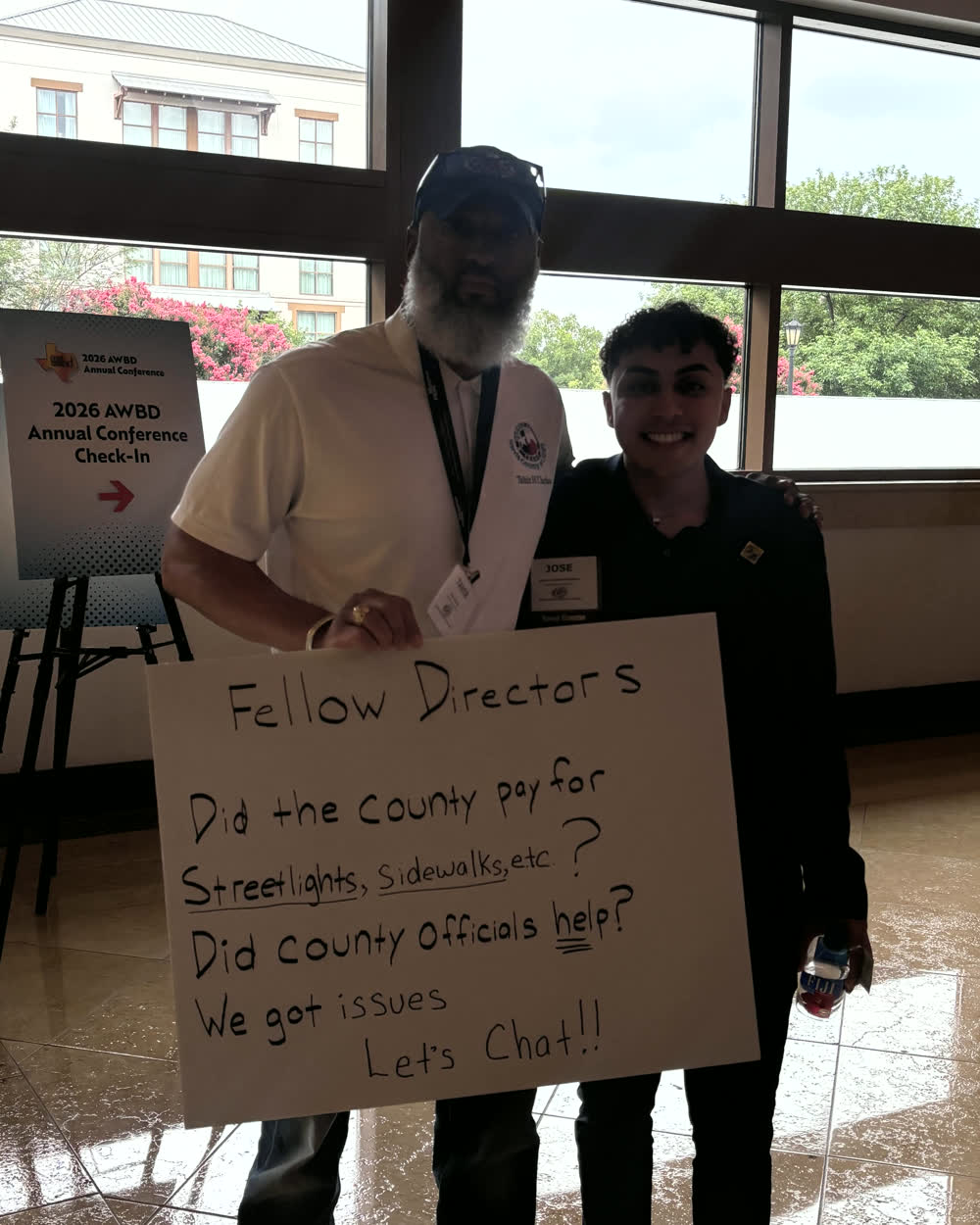 Tahir Charles at the 2026 AWBD Annual Conference holding a hand-lettered sign that reads: Fellow Directors, did the county pay for streetlights, sidewalks, etc? Did county officials help? We got issues. Let's chat.