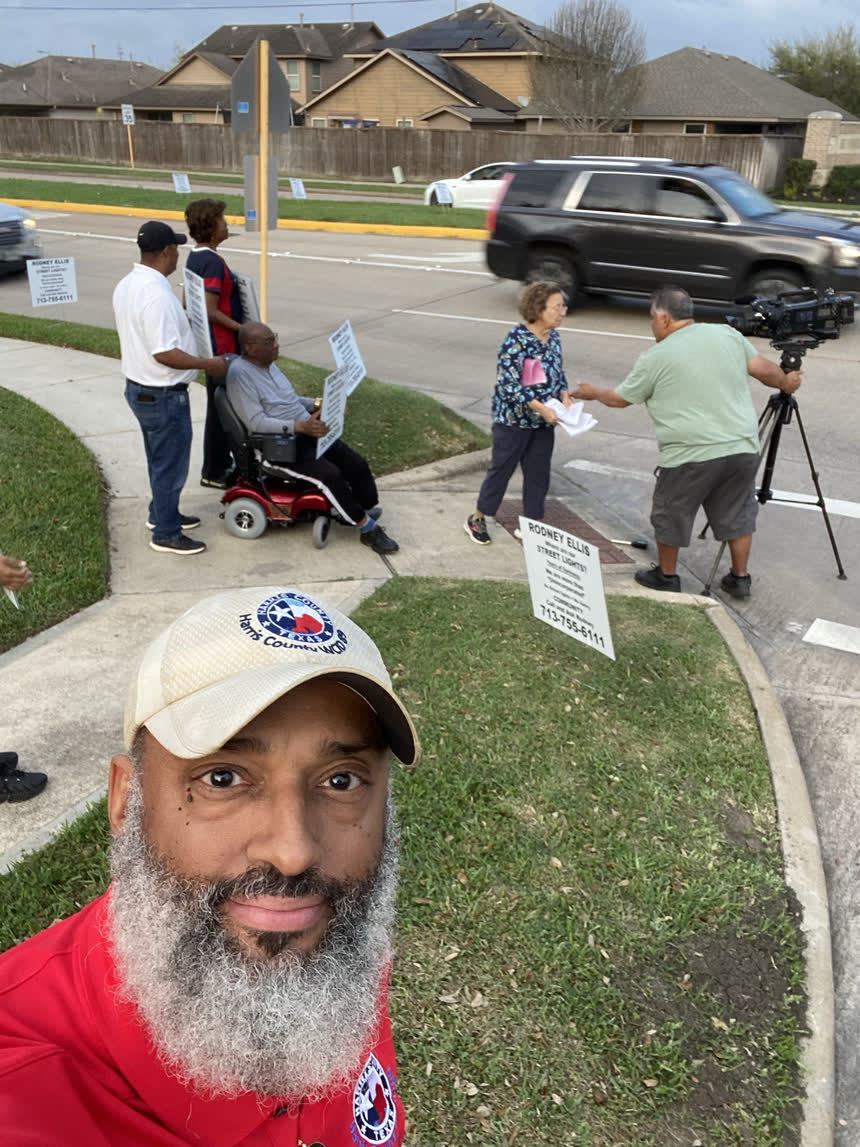 Neighbours holding “Where are our street lights?” signs on a street corner, one seated in a power wheelchair, while a resident hands papers to a television cameraman.