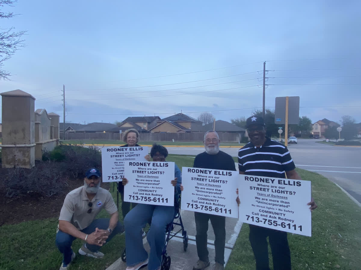 Tahir Charles kneeling beside four neighbours holding “Where are our street lights?” signs at dusk by the subdivision entrance.