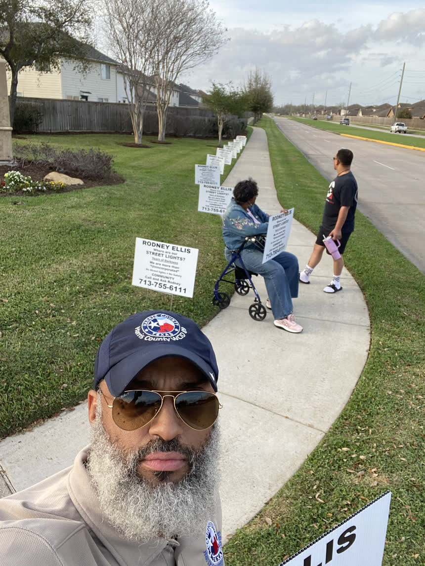 Tahir Charles in a Harris County WCID 89 cap, with a line of “Where are our street lights?” signs running down the esplanade behind him and neighbours standing along the sidewalk.