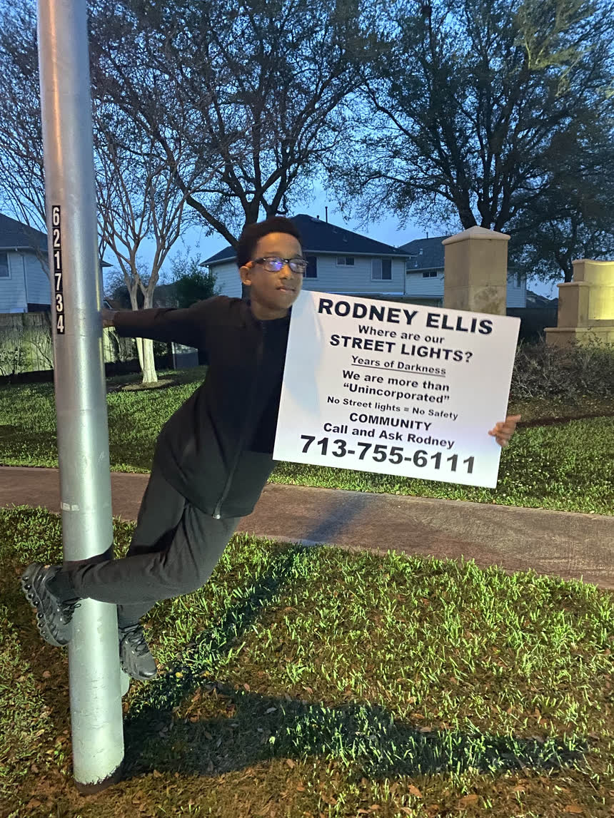 A young resident holding a “Where are our street lights?” sign while leaning off a pole.