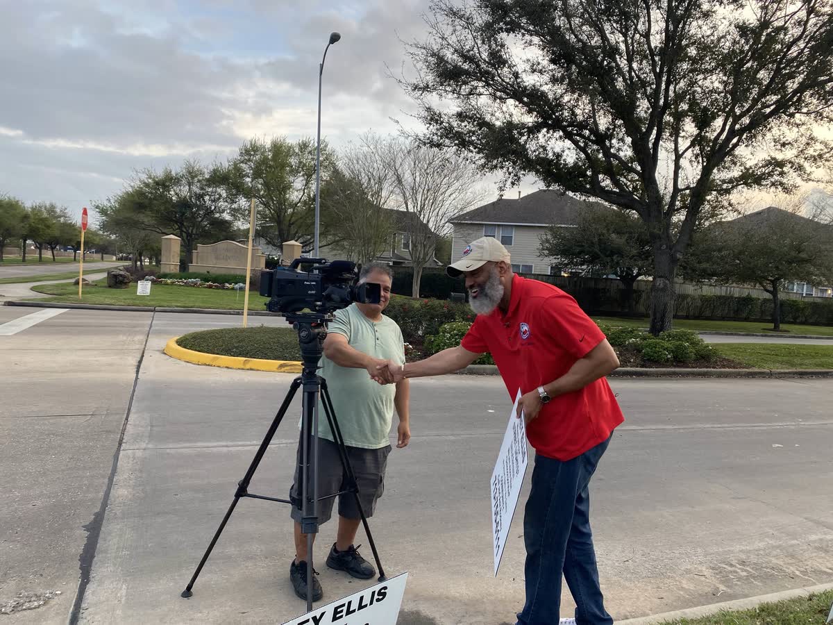 Tahir Charles shaking hands with a television cameraman standing beside his camera and tripod at the subdivision entrance.