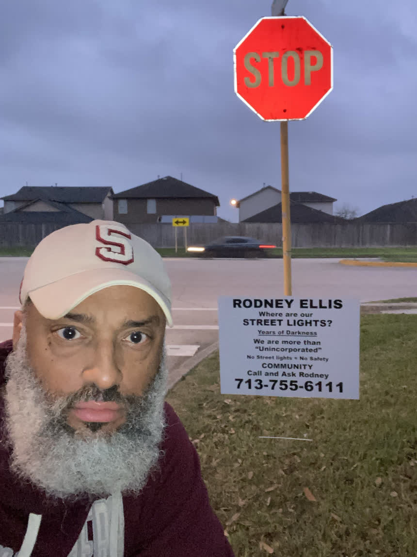 Tahir Charles beside a “Where are our street lights?” sign planted at a stop sign at dusk, with a car passing behind.