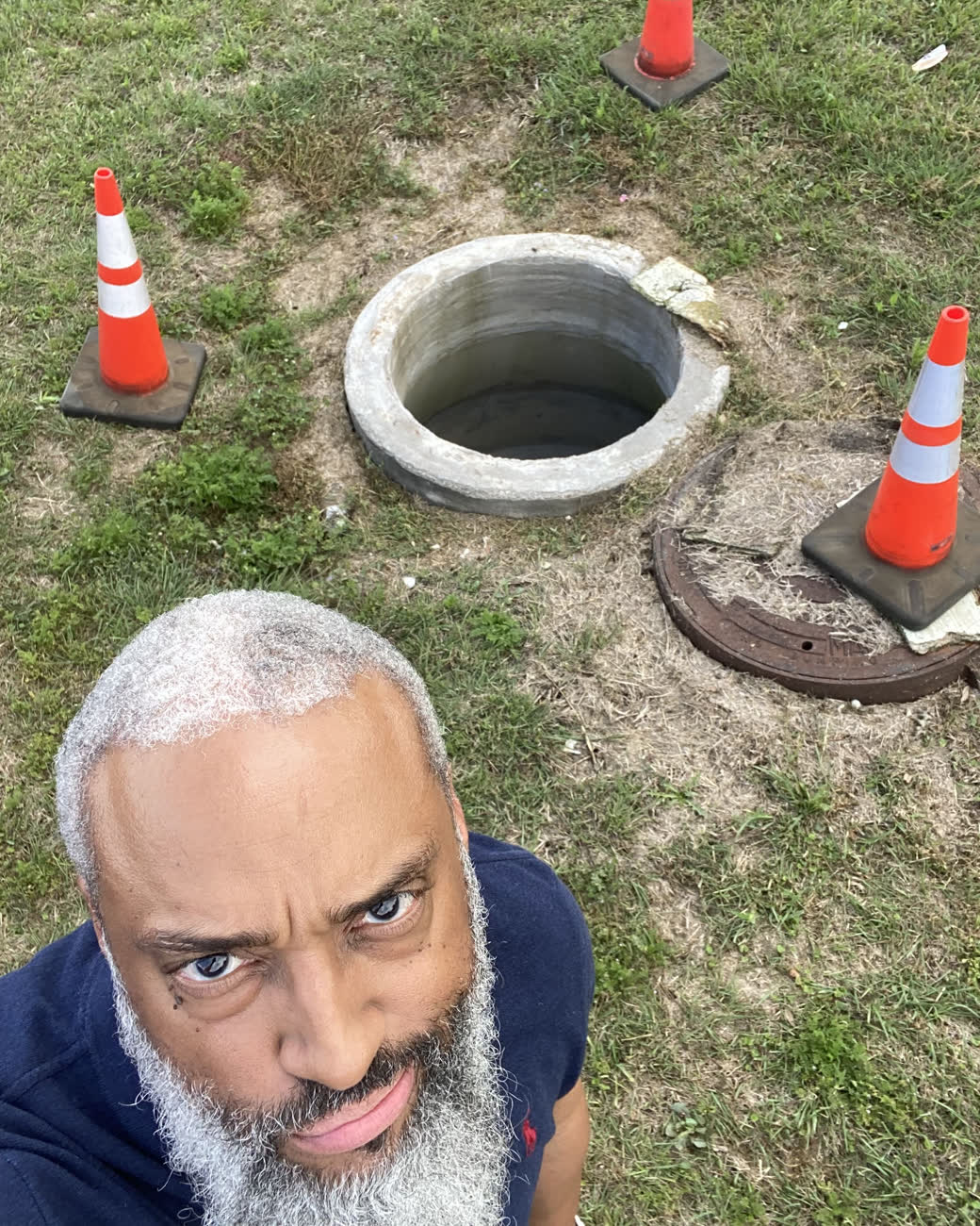 Tahir Charles standing beside an open manhole on a residential lawn, coned off with four orange traffic cones.