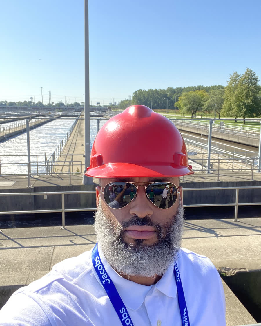 Tahir Charles in a red hard hat and safety badge, standing on a walkway above the aeration basins of a wastewater treatment plant.