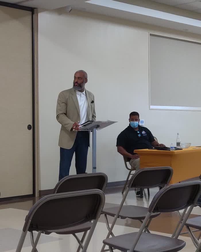 Tahir Charles speaking from a lectern at a district meeting in a community room.