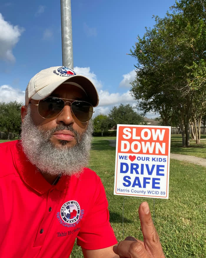 Tahir Charles pointing at a Harris County WCID 89 yard sign reading Slow Down, We Love Our Kids, Drive Safe.